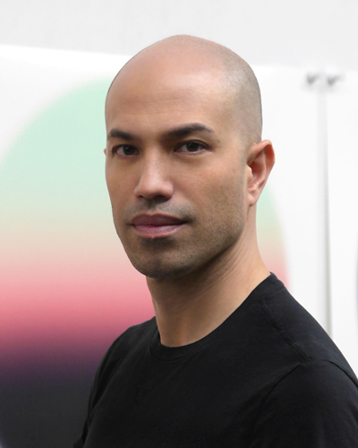 Photograph of Greg Foley in front of work, he is bald and wears a black shirt.