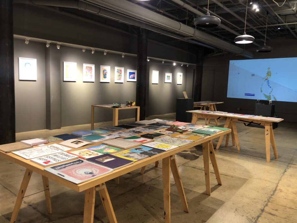 Photograph of Printing the Future Exhibition, riso prints line the walls and zines and books are arranged on wooden tables. A riso animation is projected on the back wall of the room.