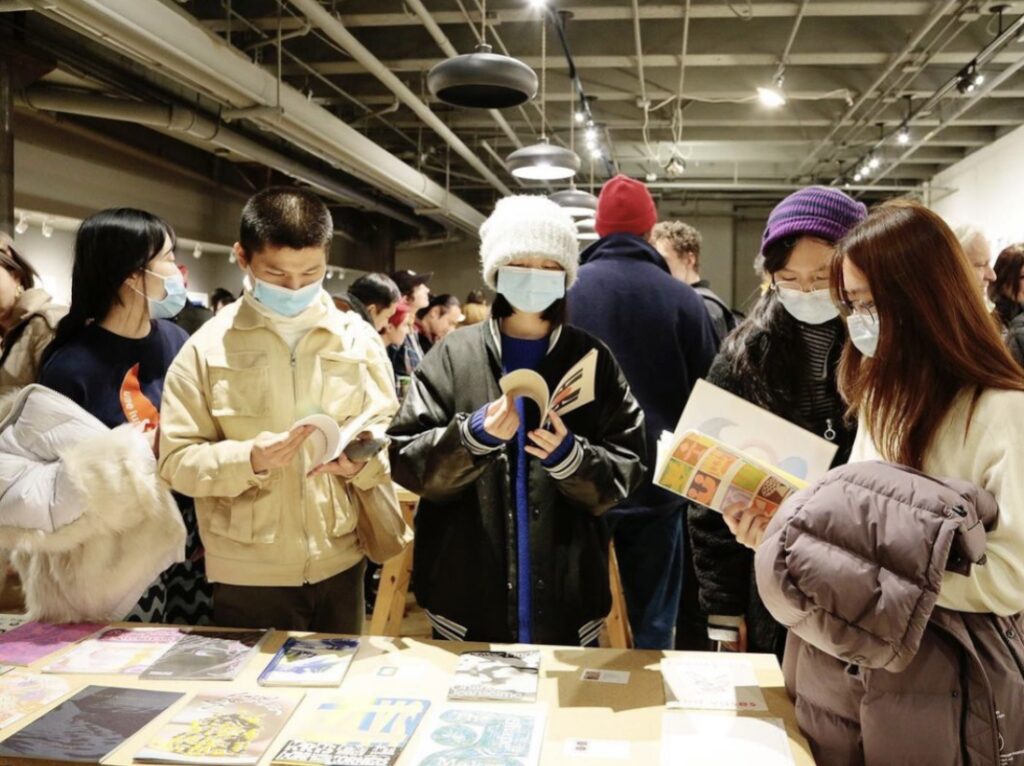 Photograph of Printing the Future opening night, many people read zines and books arranged on wooden tables.