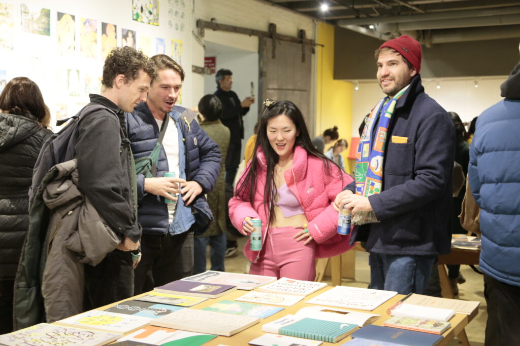 Photo of opening night at "Printing the Future" exhibition, showing zines laid out across multiple wooden tables and prints arranged on the walls salon style. Many people crowd around looking at the artwork.