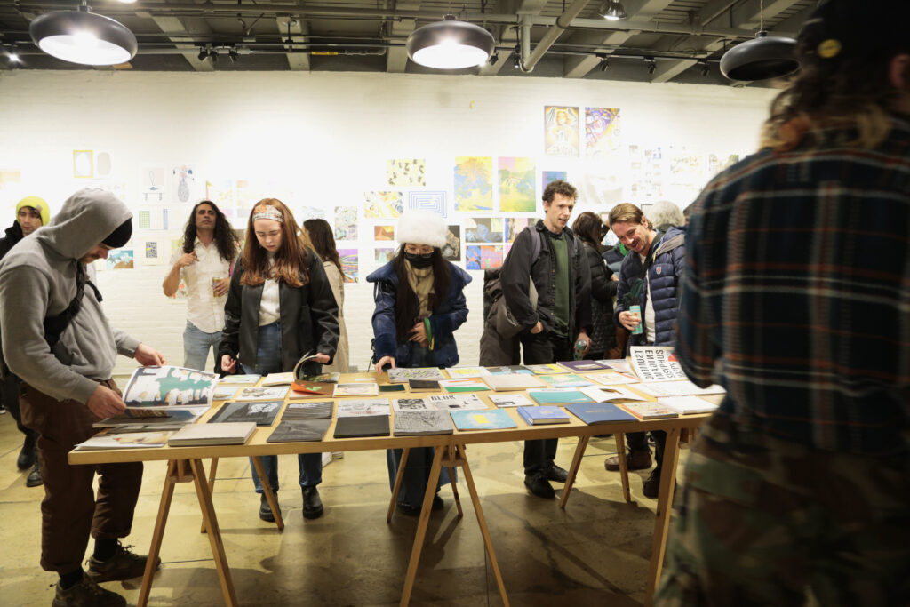 Photo of opening night at "Printing the Future" exhibition, showing zines laid out across multiple wooden tables and prints arranged on the walls salon style. Many people crowd around looking at the artwork.