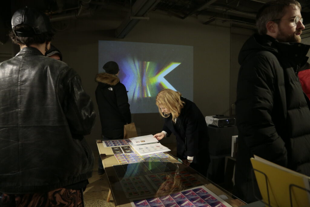 Photo of opening night at "Printing the Future" exhibition, showing zines laid out across multiple wooden tables and prints arranged on the walls salon style. Many people crowd around looking at the artwork.