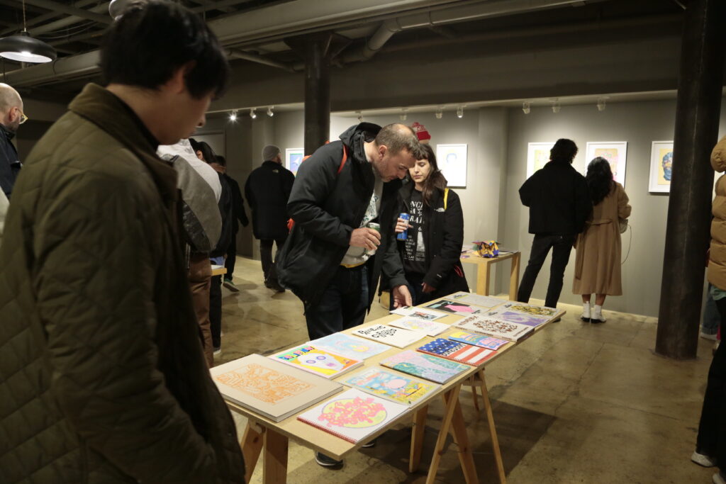 Photo of opening night at "Printing the Future" exhibition, showing zines laid out across multiple wooden tables and prints arranged on the walls salon style. Many people crowd around looking at the artwork.