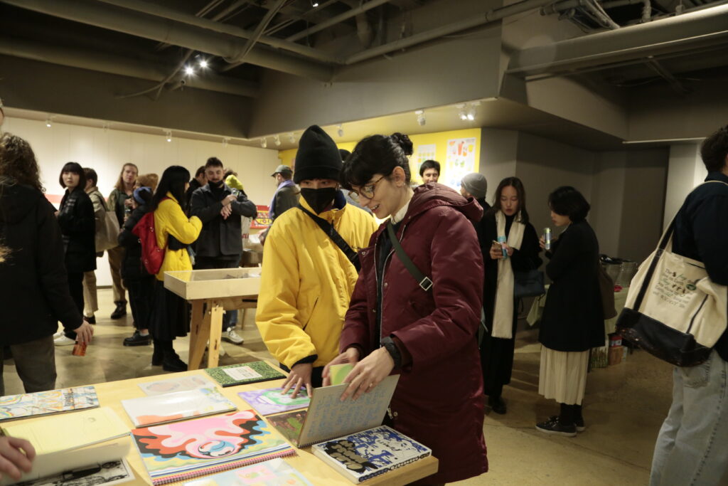 Photo of opening night at "Printing the Future" exhibition, showing zines laid out across multiple wooden tables and prints arranged on the walls salon style. Many people crowd around looking at the artwork.