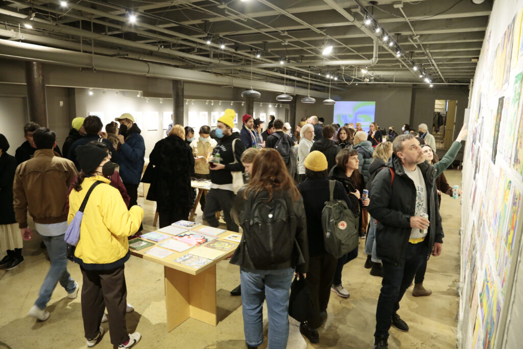 Photo of opening night at "Printing the Future" exhibition, showing zines laid out across multiple wooden tables and prints arranged on the walls salon style. Many people crowd around looking at the artwork.