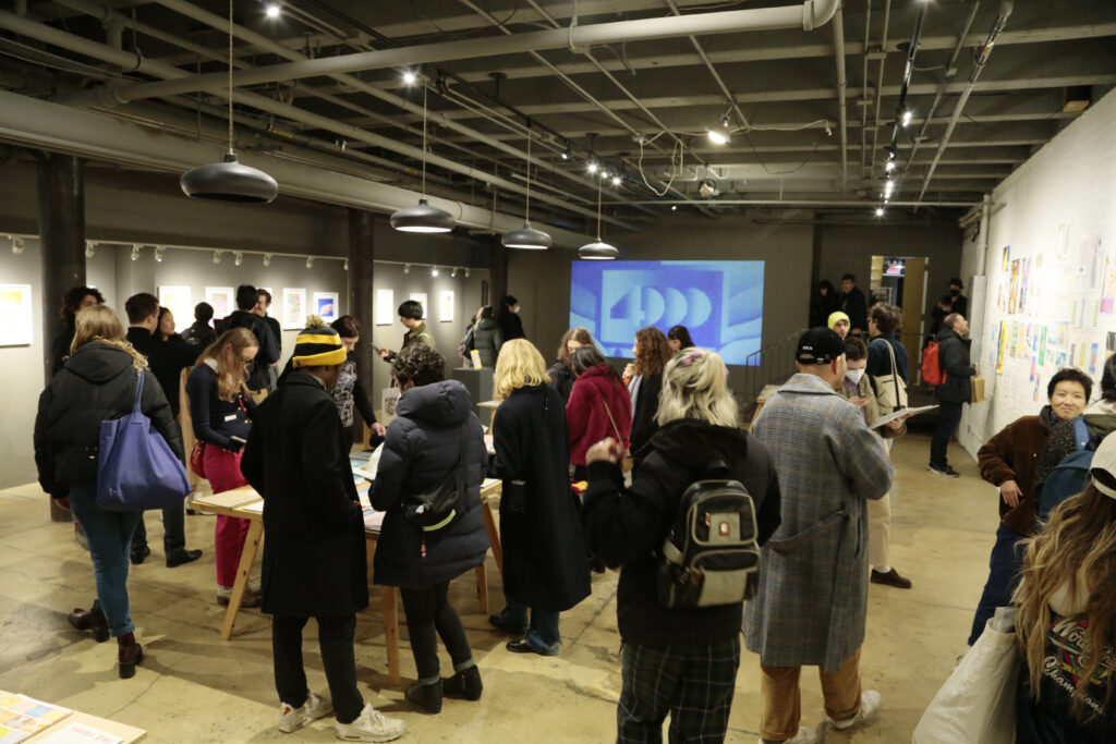 Photo of opening night at "Printing the Future" exhibition, showing zines laid out across multiple wooden tables and prints arranged on the walls salon style. Many people crowd around looking at the artwork.